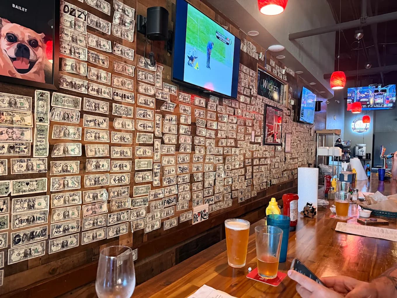 The Wicked Wall covered in dollar bills, with framed portraits and TVs above the bar